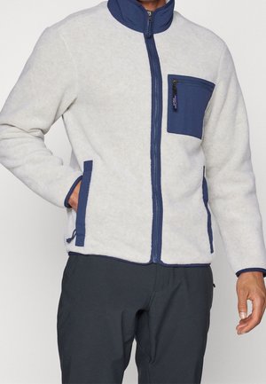 Homme portant une veste polaire blanche avec une fermeture éclair, un col, une poche et des finitions bleu marine, mains dans les poches de la veste, associée à un pantalon foncé.