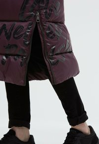 Burgundy padded jacket with glossy black text, zip closure, and a rounded hem. Paired with black jeans and black sneakers, showing rolled cuffs.