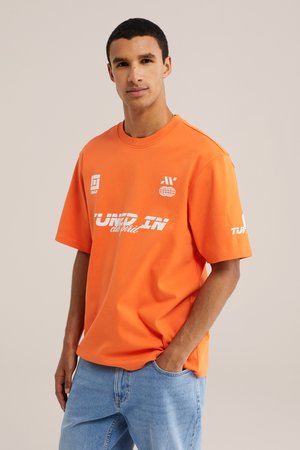 Man draagt een oranje oversized T-shirt met korte mouwen met witte logo's en lichtblauwe spijkerbroek, staat met één hand in de zak.