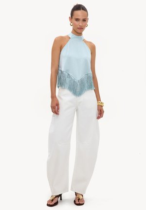 Femme portant un top à franges sans manches bleu clair, un pantalon blanc à jambes larges, des boucles d’oreilles créoles dorées, des bracelets et des sandales noires à talons hauts avec un détail floral.
