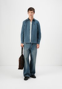 Blue button-down shirt over a white T-shirt, paired with wide-leg light blue jeans. Holding a brown suede bag, wearing black shoes.