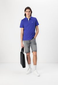 Lilla polo-shirt med hvid krave, grå denim shorts, hvide sneakers og en sort totebag. Designet er afslappet og tætsiddende.