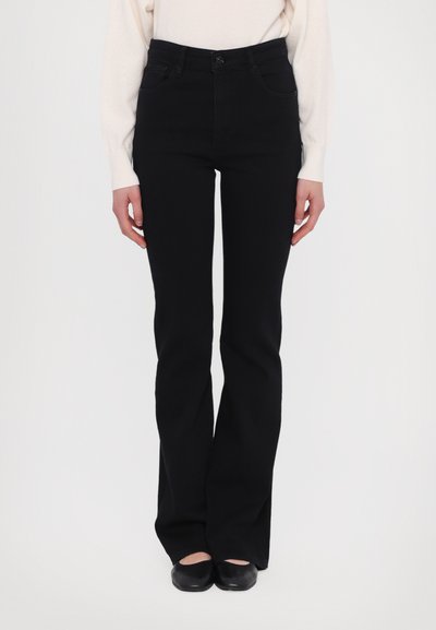Jeans évasés noirs à taille haute, cinq poches et zip à l'avant. Fabriqués en denim à la texture lisse. Portés avec un haut de couleur claire.