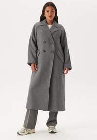 Cappotto grigio in lana doppiopetto con ampi revers, grandi bottoni e tasche laterali. Silhouette lunga e strutturata con una texture morbida.