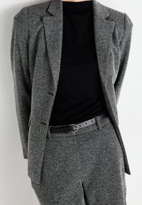 Grå texturerad kavaj med matchande byxor. Kavajen har axelvaddar och en skräddarsydd passform. Bärs över en svart skjorta med ett svart bälte som accent.