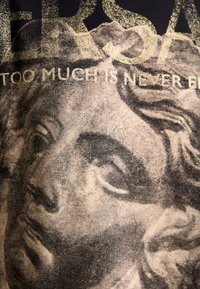 Grafische t-shirt met een textuurprint van het gezicht van een klassieke sculptuur in zwart en goud, met de tekst "TE VEEL IS NOOIT GENOEG."