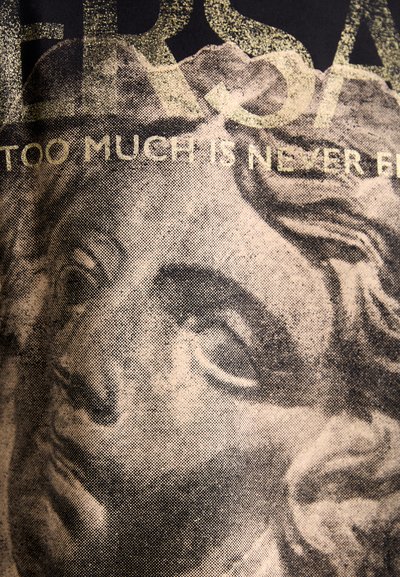 T-shirt graphique avec une impression texturée du visage d'une sculpture classique en noir et or, accompagné du texte "TROP N'EST JAMAIS ASSEZ."