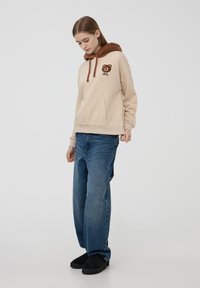 Felpa beige con cappuccio in finta pelliccia marrone, caratterizzata da una grafica di un orso e lacci marroni. Abbinata a jeans blu e scarpe nere senza lacci.