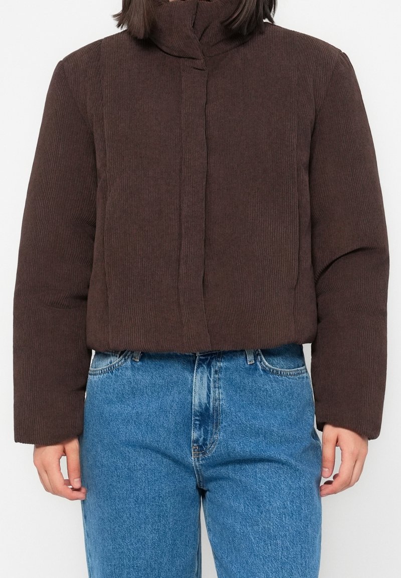 Personne portant une veste matelassée en velours côtelé marron avec un col montant et un jean bleu en denim, debout, les bras détendus le long du corps.