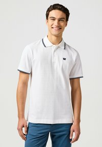 Weißes Poloshirt mit strukturierter Oberfläche, dunkelblauem gestreiften Kragen und kleinem Logo. Kurzärmlig und mit geradem Saum, kombiniert mit blauen Hosen.