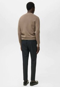 Man staat met de rug naar de camera gekeerd, gekleed in een beige trui, donkere op maat gemaakte broek en zwarte leren instapschoenen, tegen een effen lichte achtergrond.