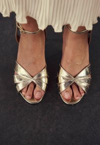 Metalliske guld sandaler har et krydsforbindelsesdesign og åben tå. Stropperne er slanke med en glat overflade og en let glans.