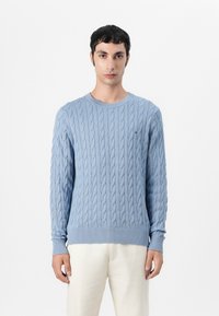 Pulover din lână cu un model tricotat în culori albastre deschise, cu decolteu rotund și manșete cu model în care. Prezintă un logo mic pe piept. Textură netedă și design ajustat.