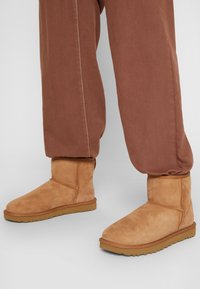 Botas de tobillo de ante color marrón claro con puntera redondeada y detalles cosidos, combinadas con pantalones marrones de corte holgado. Suela de goma texturada visible.