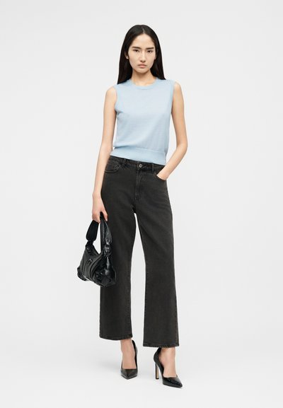 Frau trägt ein hellblaues ärmelloses Oberteil, schwarze Jeans mit weitem Bein, schwarze High Heels und hält eine schwarze Handtasche, während sie eine Hand in der Tasche hat.