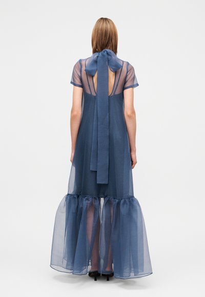 Γυναίκα που φορά ένα διάφανο, ναυτικό μπλε φόρεμα με φιόγκο στον λαιμό και μακριά φούστα με βολάν, όρθια με την πλάτη της να κοιτάζει προς τον θεατή.