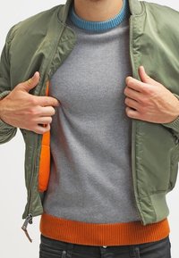 Grüne Bomberjacke über einem grauen Pullover mit oranger Rippborte. Die Jacke hat einen blauen Kragen und verfügt über ein oranges Innenfutter.
