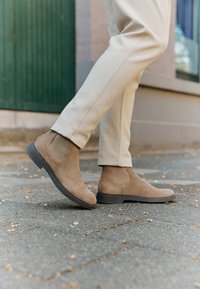 Botas chelsea de ante en marrón claro con paneles elásticos laterales y suela de goma negra, combinadas con pantalones beis ajustados en una acera.