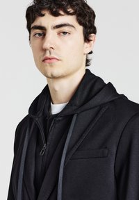 Schwarze, doppellagige Jacke mit kapuze zum Zipper, kontrastierenden Kordeln und einer Brusttasche, mit einer glatten, matten Textur.