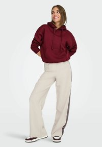 Felpa cotone bordeaux con tasca frontale, abbinata a pantaloni a gamba larga beige con strisce laterali nere e scarpe bianche.