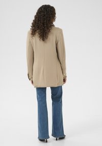 Beige dlouhý blazer s přiléhavým střihem, se knoflíky na manžetách, kombinovaný s modrými rozšířenými džíny a černými botami na podpatku.
