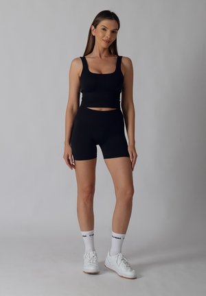 Woman standing wearing black sleeveless crop top and biker shorts, white socks, and white sneakers against plain gray background.