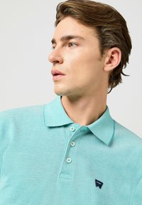 Polo shirt turchese in tessuto testurizzato. Presenta un colletto, tre bottoni e un piccolo logo navy sul lato sinistro del petto.