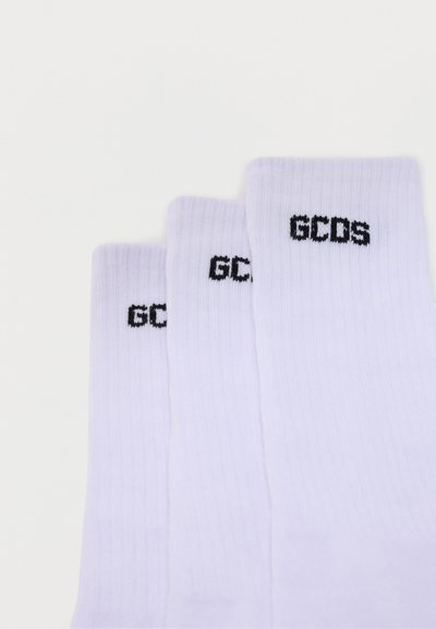 Trois chaussettes blanches côtelées montantes avec le logo GCDS noir brodé près du haut de la bande, disposées en chevauchement sur un fond blanc.
