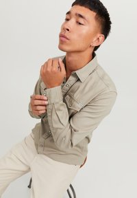 Camisa de botón color beige claro con mangas largas, botones a presión y bolsillo en el pecho. Tejido suave con un corte casual. Modelo sentado.