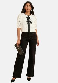 Blusa bianca in pizzo con maniche a palloncino, dettaglio in nastro nero, abbinata a jeans neri a vita alta, mentre tiene una borsa a mano nera, stilizzata con tacchi a punta.