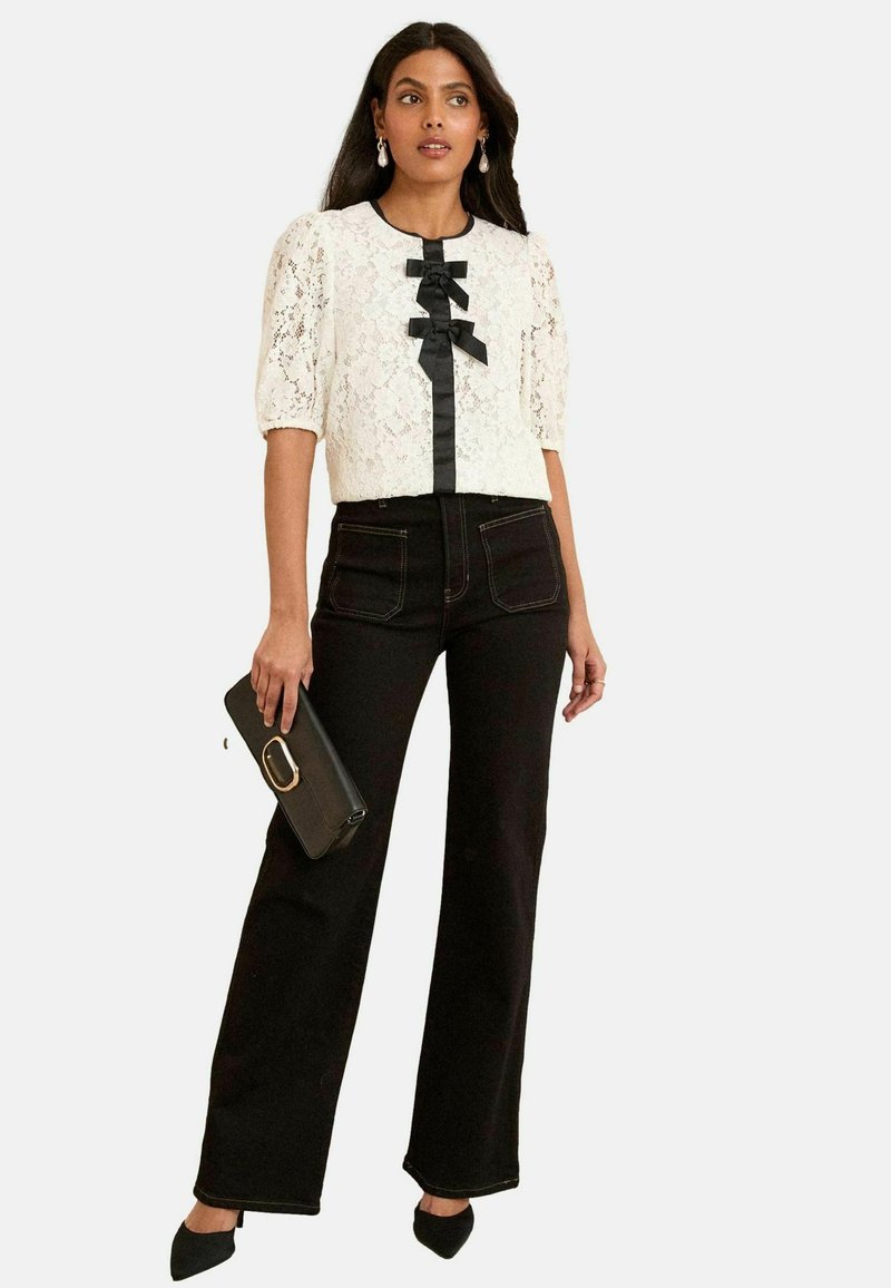Blusa bianca in pizzo con maniche a palloncino, dettaglio in nastro nero, abbinata a jeans neri a vita alta, mentre tiene una borsa a mano nera, stilizzata con tacchi a punta.