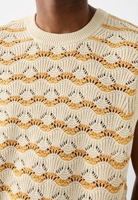 Maglione senza maniche color crema con motivi ondulati alternati in oro e marrone, caratterizzato da dettagli a traforo e un colletto a coste.