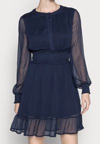 Marinblå klänning med långa genomskinliga ärmar, knäppning framtill, elastisk midja och flerskiktad volangkant. Mjukt, lätt tyg.