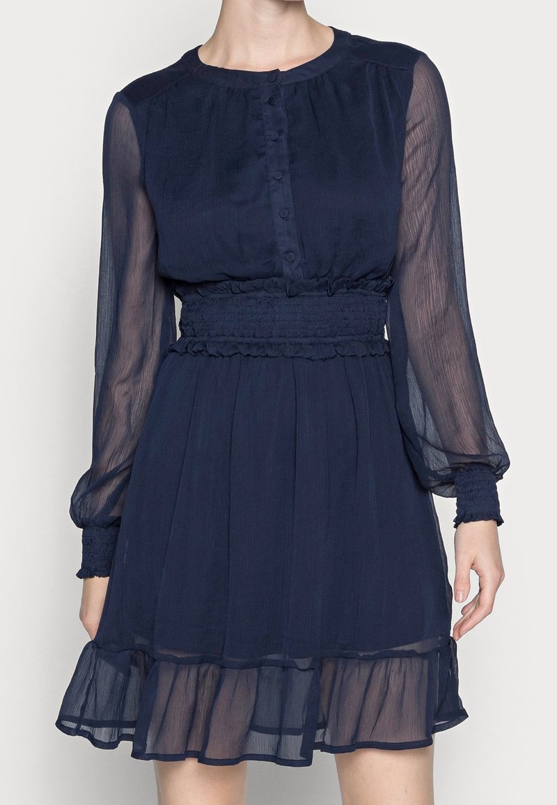 Marinblå klänning med långa genomskinliga ärmar, knäppning framtill, elastisk midja och flerskiktad volangkant. Mjukt, lätt tyg.