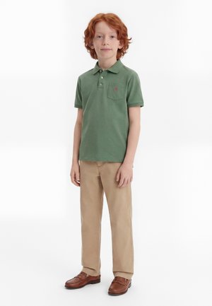 Rødharret dreng står iført en grøn polo-shirt, beige bukser og brune loafers mod en ensfarvet hvid baggrund.