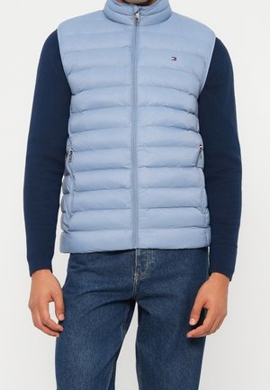 Homme portant une veste matelassée bleu clair zippée sur une chemise bleu foncé à manches longues et un jean bleu, debout avec les bras détendus le long du corps.