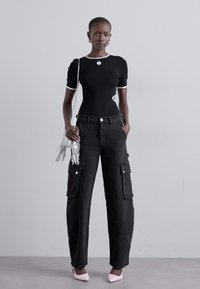 Top negru ajustat cu margine albă, pantaloni cargo negri cu buzunare mari, tocuri roz, și o geantă de umăr albă cu franjuri. Fundal simplu.