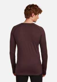 Man met krullend rood haar, gekleed in een aansluitend, donkerpaars shirt met lange mouwen, gezien van achteren tegen een effen lichte achtergrond.