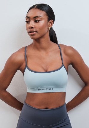 Femme aux cheveux foncés attachés en queue de cheval, portant un soutien-gorge de sport bleu clair et des leggings gris, regardant sur le côté avec les mains dans le dos.