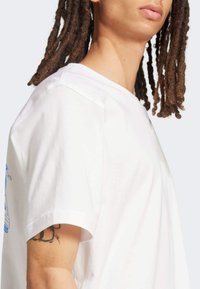 Bijela pamučna majica s okruglim izrezom, kratkim rukavima i plavim grafičkim dizajnom s lijeve strane. Glatka tekstura, ležeran kroj.