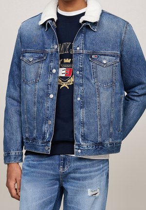 Muž v modré džínové bundě s bílým límcem z ovčí vlny, tmavě modré mikině s logem a světle modrých roztrhaných džínách, stojící s rukou v kapse.