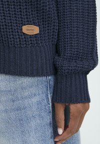 Stickad marinblå tröja med ett mönster av strukturerad stygn, ribbade ärmslut och en brun läderetikett med varumärket på kanten.