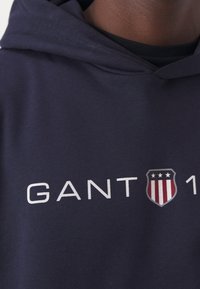 Persona vestida con una sudadera con capucha azul marino que lleva en el pecho "GANT" y un logotipo de escudo con estrellas y rayas en rojo, blanco y azul.