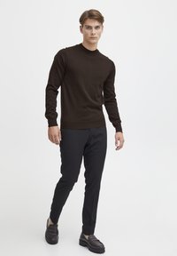 Maglione marrone a maniche lunghe con scollatura a girocollo, abbinato a pantaloni neri su misura e mocassini scuri, in piedi su uno sfondo semplice.