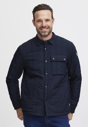 Navy quilted jacket with a button front, pointed collar, and a pocket on the left chest. Features textured fabric and subtle stitching detail.