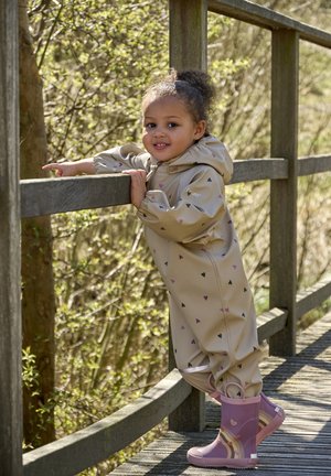 Mono impermeable beige con patrones de corazones en negro y morado. Botas de goma moradas con diseños. Niño apoyado en una barandilla de madera.