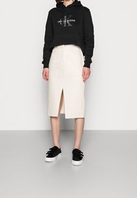 Persona vistiendo un hoodie corto negro y una falda midi color crema con una abertura frontal, combinada con zapatillas negras, se encuentra de pie sobre un fondo neutro.