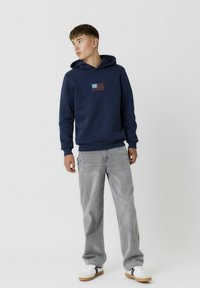 Marineblauer Kapuzenpullover mit einem vorderen Grafikumdruck einer kleinen Flagge, kombiniert mit hellgrauen Jeans und weißen Sneakern mit schwarzen Akzenten.