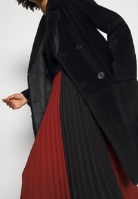 Svart fuskpäls dubbelknäppt kappa över en svart och röd plisserad kjol. Kjolen har vertikala plisseringar och två färgblock.