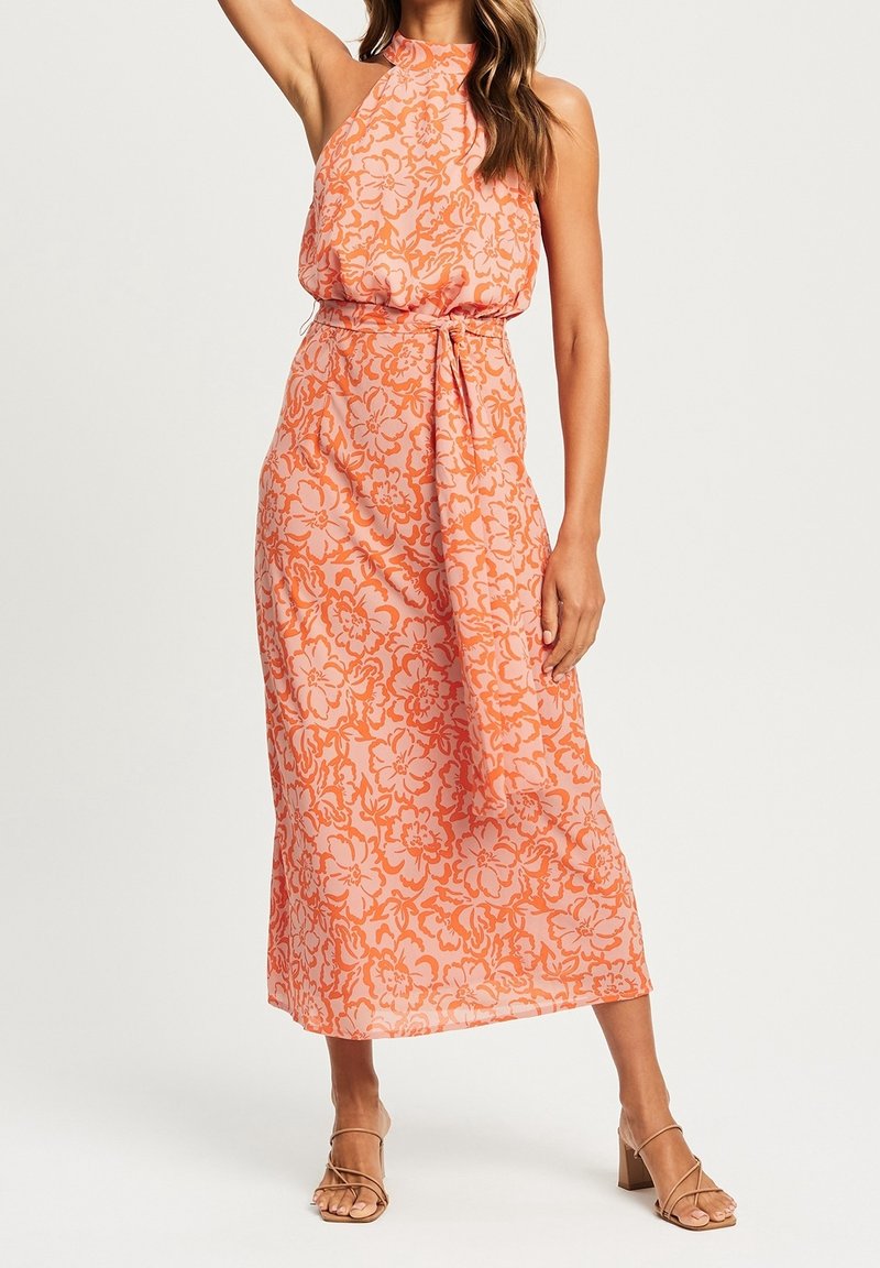 Femme portant une robe midi orange sans manches à col haut, ornée de motifs floraux, avec une ceinture nouée à la taille et des sandales à talons ouvertes couleur beige.